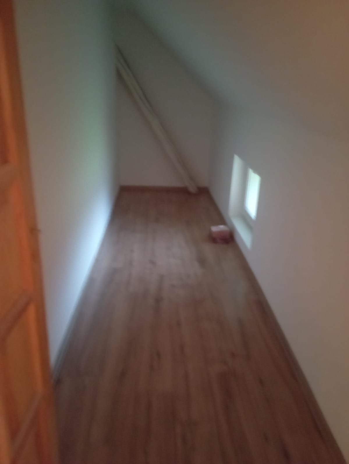 Poddasze z drewnianą podłogą, skośnym sufitem i małym oknem, widoczna rura pod sufitem, niedokończone wnętrze.