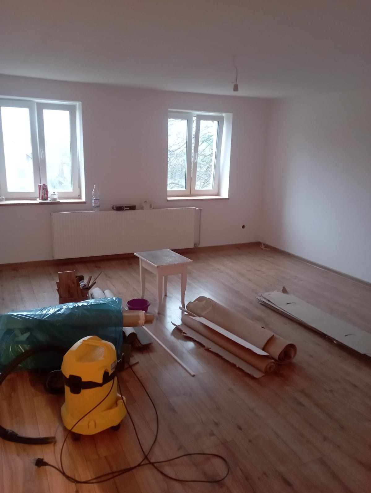 Pokój po malowaniu ścian na biało, z drewnianą podłogą, dwoma oknami, grzejnikiem, małym stolikiem, workiem na śmieci, odkurzaczem i rozłożonymi rolkami papieru zabezpieczającego.