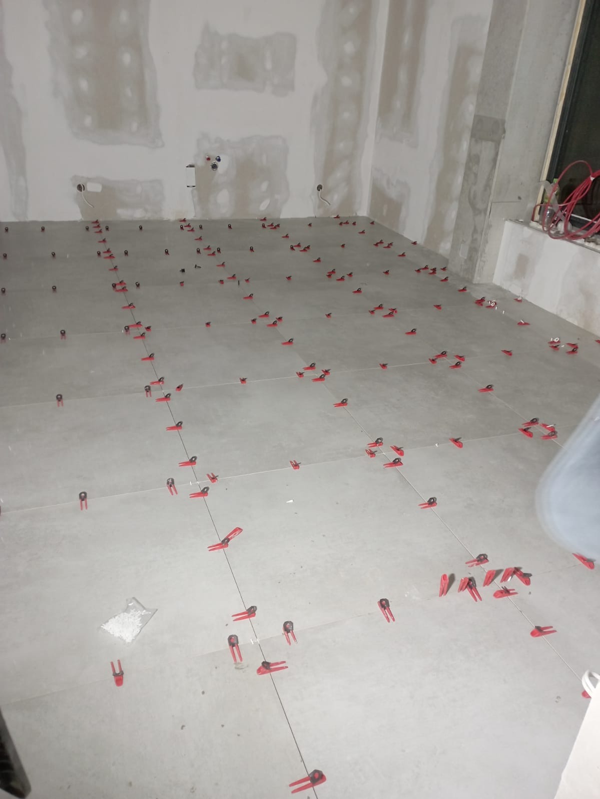 Szare płytki podłogowe w trakcie układania, z widocznymi czerwonymi klipsami poziomującymi i sznurkiem wyznaczającym linie, ściany przygotowane pod wykończenie.