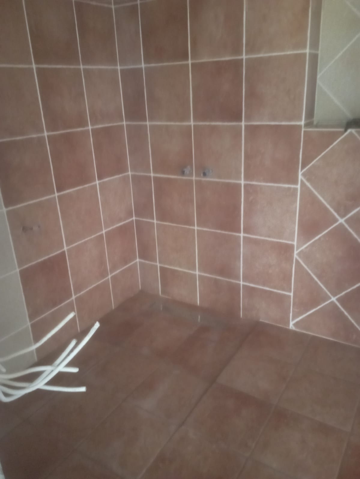 Brązowe płytki ścienne i podłogowe w surowej łazience, widoczne wystające rury instalacyjne, brak armatury.