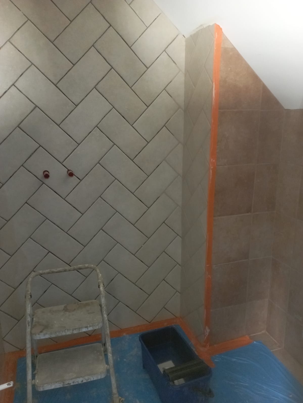 Ściana łazienki wyłożona jasnoszarymi płytkami w jodełkę, widoczne dwa otwory na baterię, drabina malarska i kuweta z wałkiem malarskim, zabezpieczona folią podłoga.