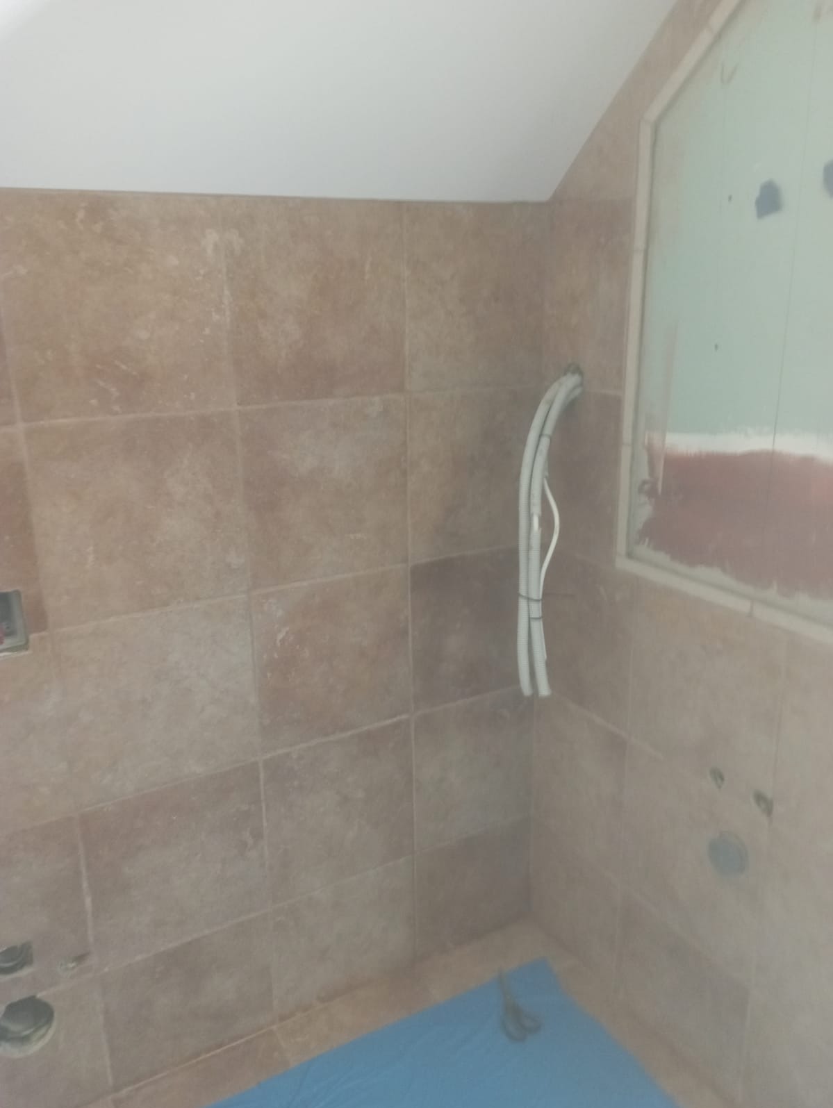 Ściana łazienki w trakcie wykańczania, z widocznymi beżowymi płytkami, wystającymi przewodami elektrycznymi zabezpieczonymi taśmą, nieotynkowanym oknem i niebieską folią ochronną na podłodze.