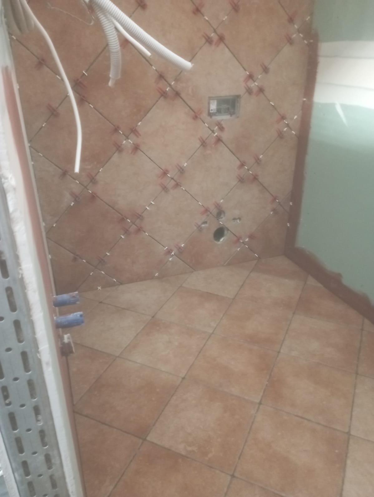 Brązowe płytki ułożone na podłodze i ścianach łazienki, z plastikowymi krzyżykami dystansowymi, widoczne przewody i otwory na instalacje.