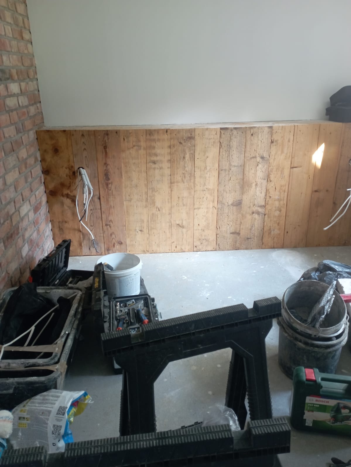 Przygotowanie do prac wykończeniowych: drewniana boazeria na ścianie, widoczne kable elektryczne, skrzynki z narzędziami Stanley i Bosch, wiadro z zaprawą i kozioł roboczy.