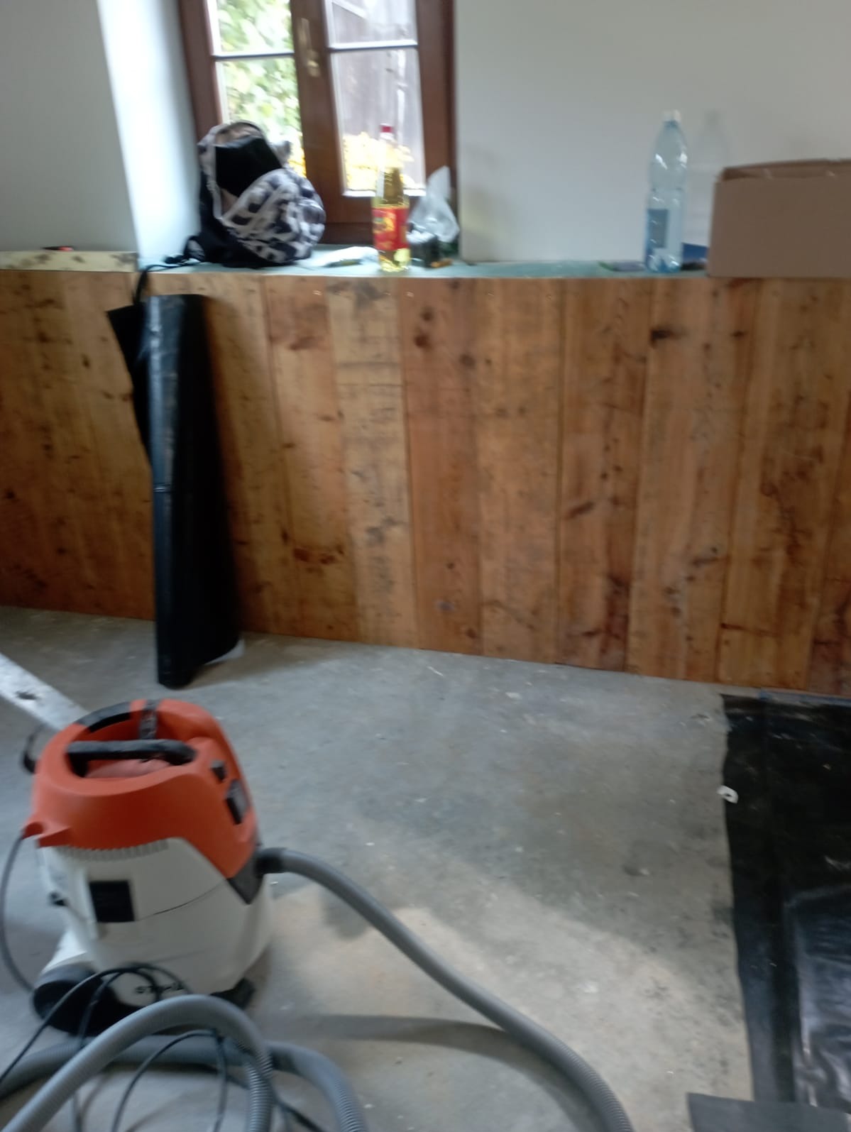 Ściana wyłożona drewnianymi panelami, widoczny odkurzacz przemysłowy, okno z plecakiem i butelkami na parapecie, oraz folia ochronna na podłodze, sugerujące prace wykończeniowe.