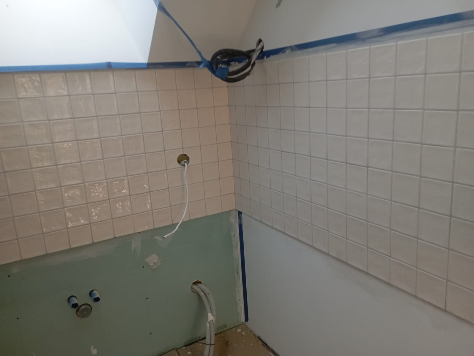 Fragment ściany łazienki w trakcie prac wykończeniowych, widoczne ułożone białe płytki, niezabezpieczona płyta gipsowo-kartonowa z instalacją hydrauliczną, taśma malarska zabezpieczająca krawędzie,...