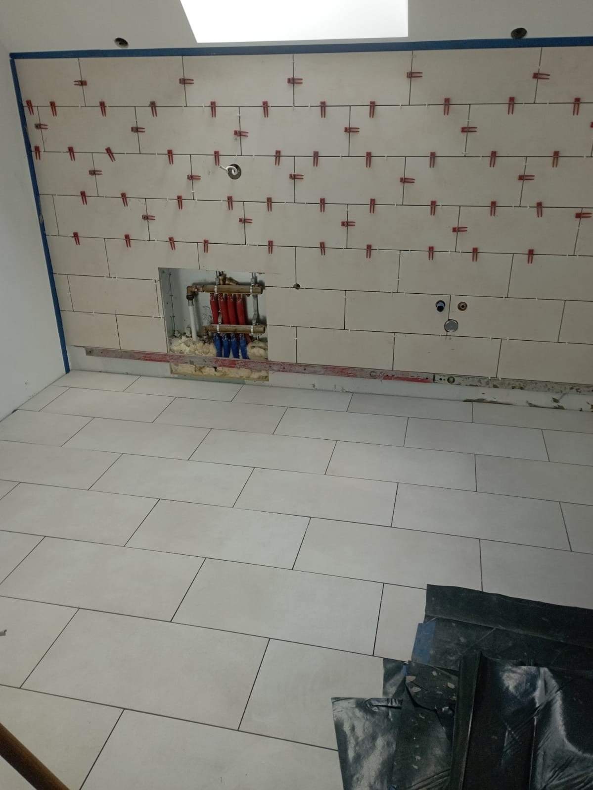 Świeżo ułożone, podłużne płytki podłogowe i częściowo wyłożona płytkami ściana w łazience, z widocznymi czerwonymi krzyżykami dystansowymi, instalacją wodną w ścianie i poziomnicą.