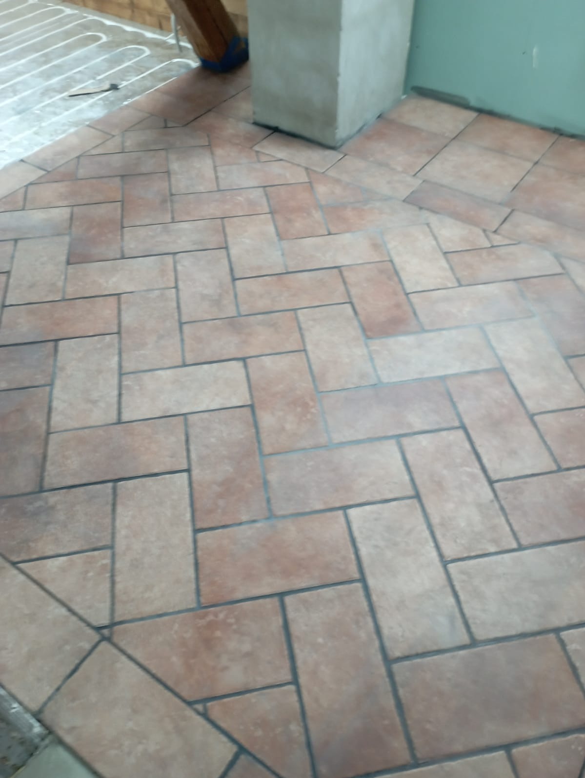 Ułożona podłoga z płytek ceramicznych w odcieniach beżu i brązu, wzór jodełki w łazience, widoczne ogrzewanie podłogowe w rogu pomieszczenia.