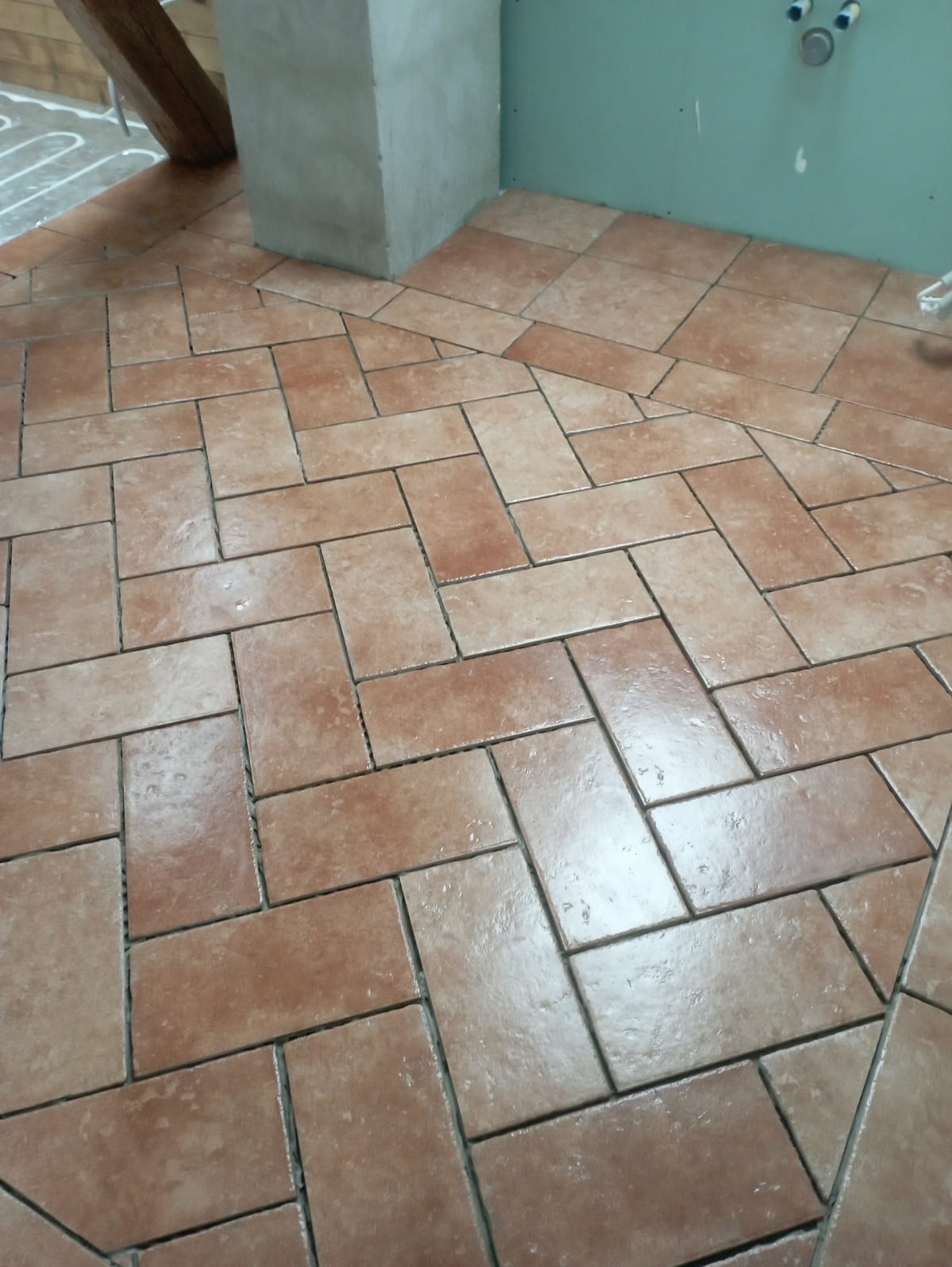 Ułożona terakota w jodełkę na podłodze łazienki w trakcie prac wykończeniowych, widoczne rury ogrzewania podłogowego i surowe ściany.