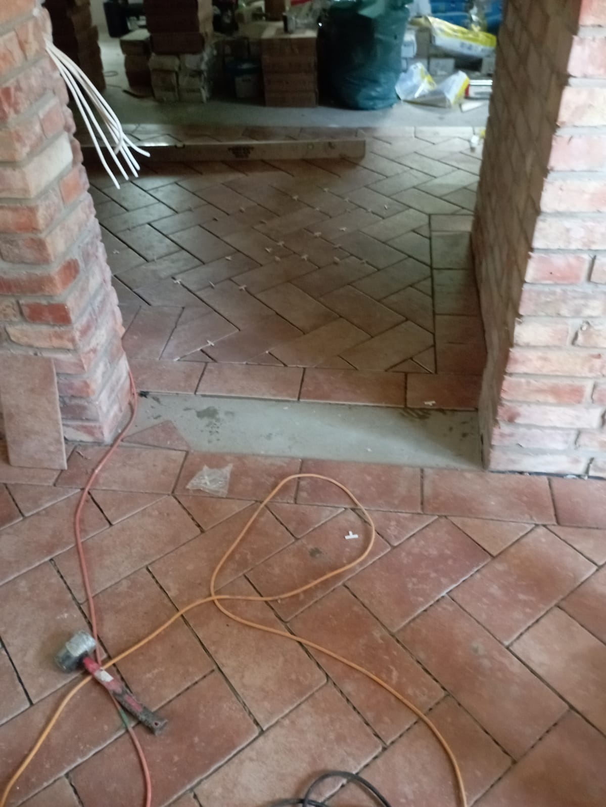 Ułożona podłoga z terakoty w jodełkę, widoczny fragment ceglanego filara, kabel elektryczny i młotek, etap prac wykończeniowych.