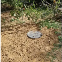 Studnia z metalową rurą i zaworami wystająca ponad ziemię, przykryta okrągłą pokrywą studzienną na piaszczystym podłożu, zarośla w tle.