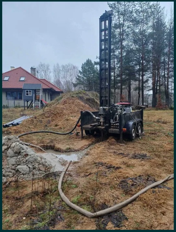 Mobilna wiertnica studni głębinowej w trakcie pracy, z widocznymi wężami i błotnistym terenem, w tle dom z czerwonym dachem i las.
