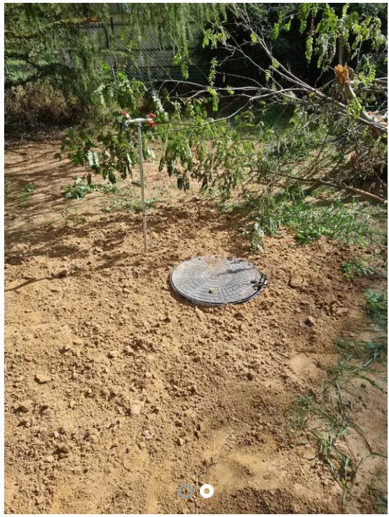 Studnia z metalową rurą i zaworami wystająca ponad ziemię, przykryta okrągłą pokrywą studzienną na piaszczystym podłożu, zarośla w tle.