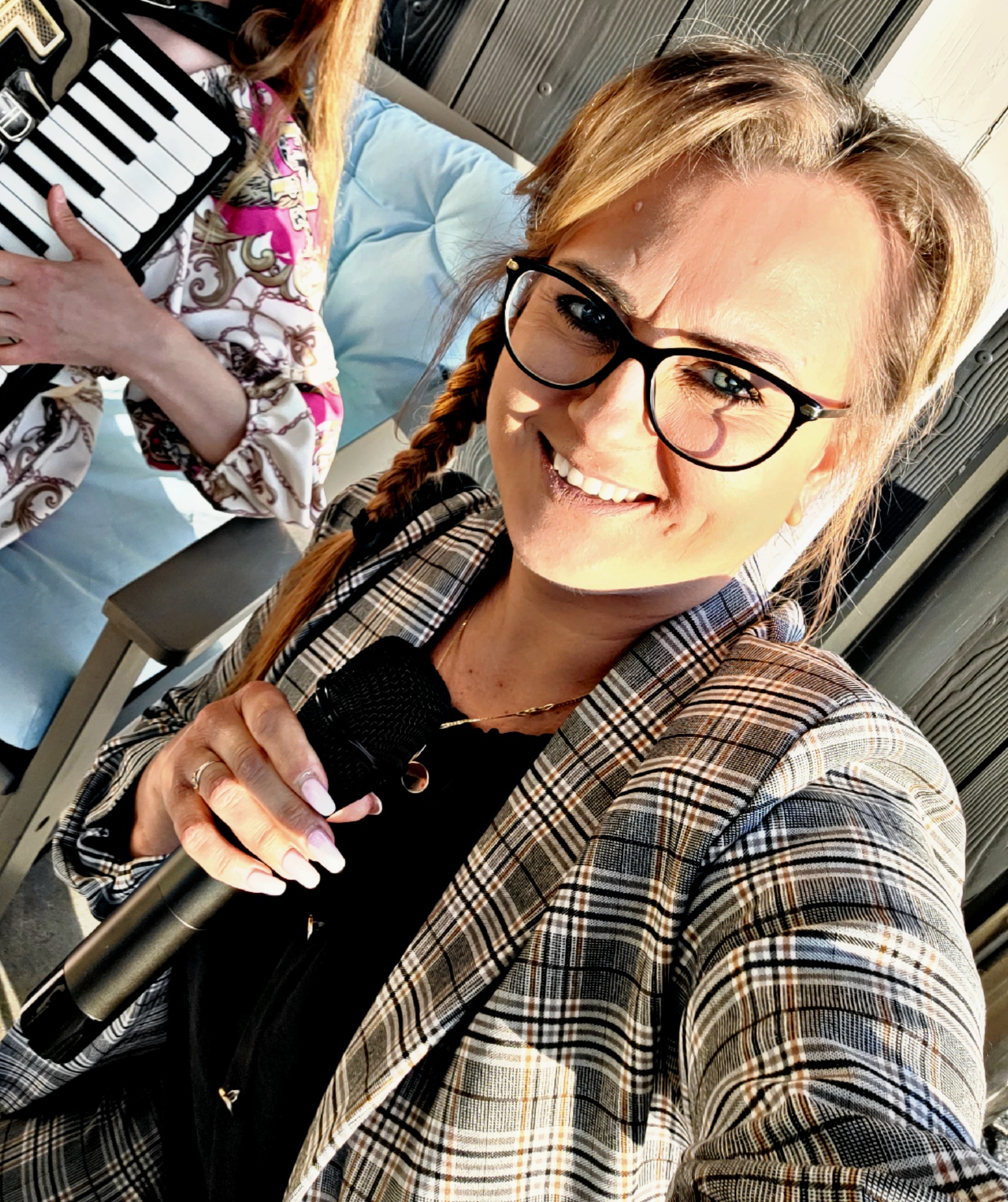 Uśmiechnięta kobieta w okularach i kraciastej marynarce trzyma mikrofon, obok widoczny fragment akordeonu i inna osoba.