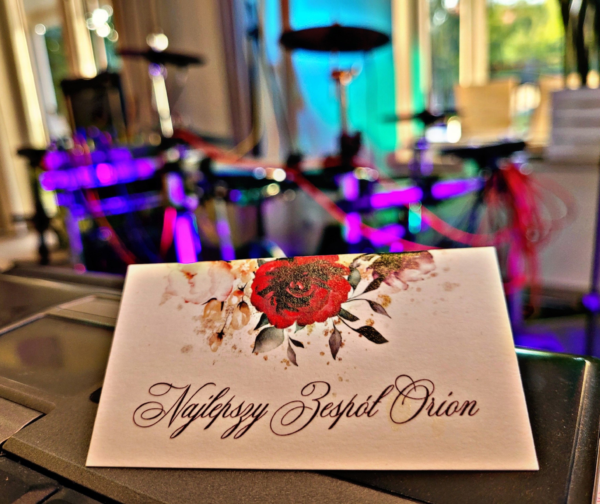 Wizytówka z napisem 'Najlepszy Zespół Orion' na tle rozmytej perkusji i kolorowych świateł scenicznych.