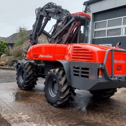 MIKOP Michał Misiek - Pomarańczowa koparka Mecalac 12MTX na mokrym betonowym podłożu, z widocznym budynkiem w tle.