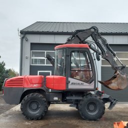 MIKOP Michał Misiek - Czerwono-szara koparka kołowa Mecalac 12MTX zaparkowana przed szarym budynkiem z bramą garażową, widok z boku na mokrym podłożu.