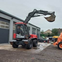MIKOP Michał Misiek - Czerwono-szara koparka kołowa Mecalac na terenie budowy, widok z przodu, operator w kabinie, lekko pochmurne niebo.