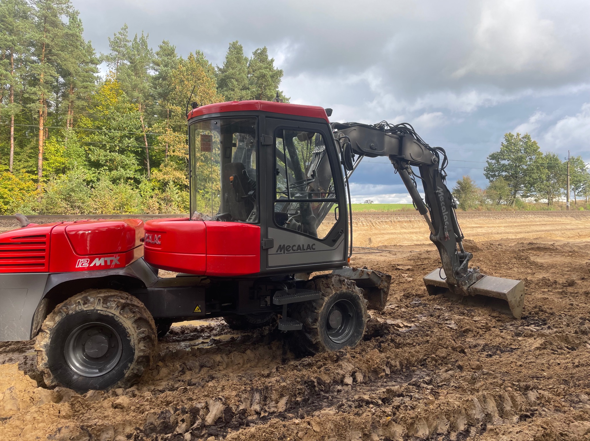 Czerwono-szara koparka kołowa Mecalac 12MTX na placu budowy, z widocznym wykopem i błotnistym terenem, las w tle pod pochmurnym niebem.
