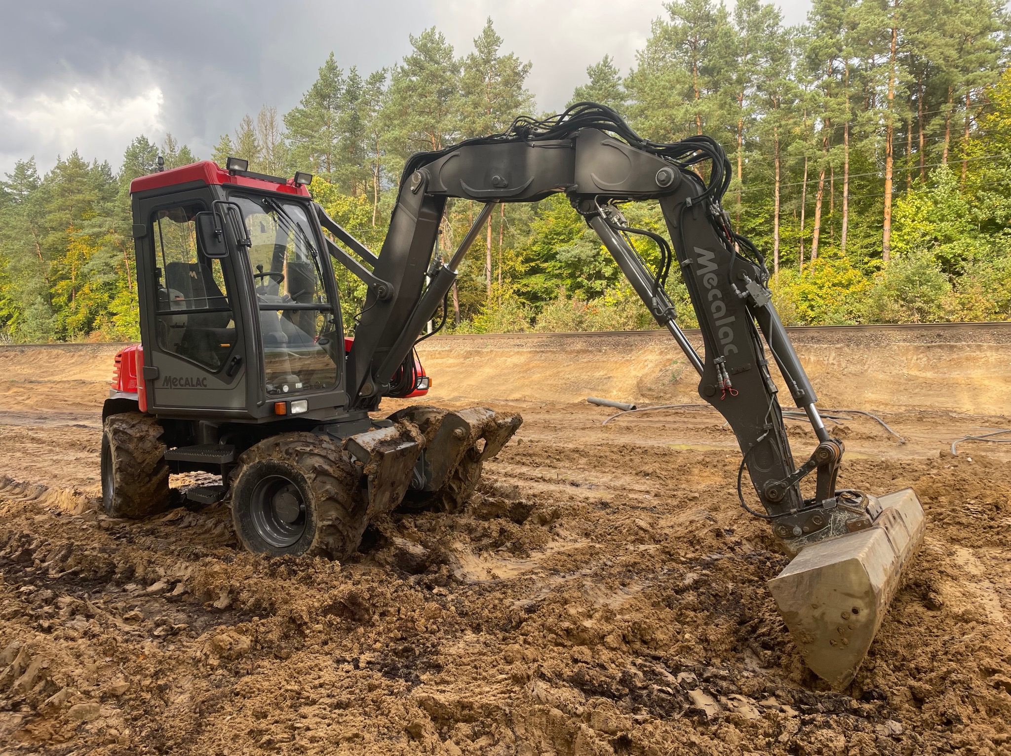 Czerwono-szara koparka kołowa Mecalac z łyżką zagłębia się w błotnistej, brązowej ziemi na tle wykopu i lasu pod pochmurnym niebem.