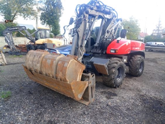 Czerwona koparka kołowa z dużą, rdzawą łyżką wieloczerpakową na nierównym, szarym terenie; w tle inne maszyny budowlane.