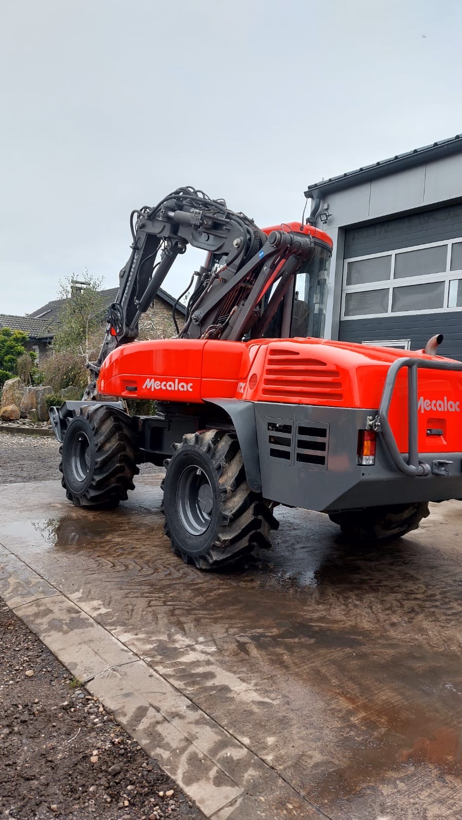 Pomarańczowa koparka Mecalac 12MTX na mokrym betonowym podłożu, z widocznym budynkiem w tle.
