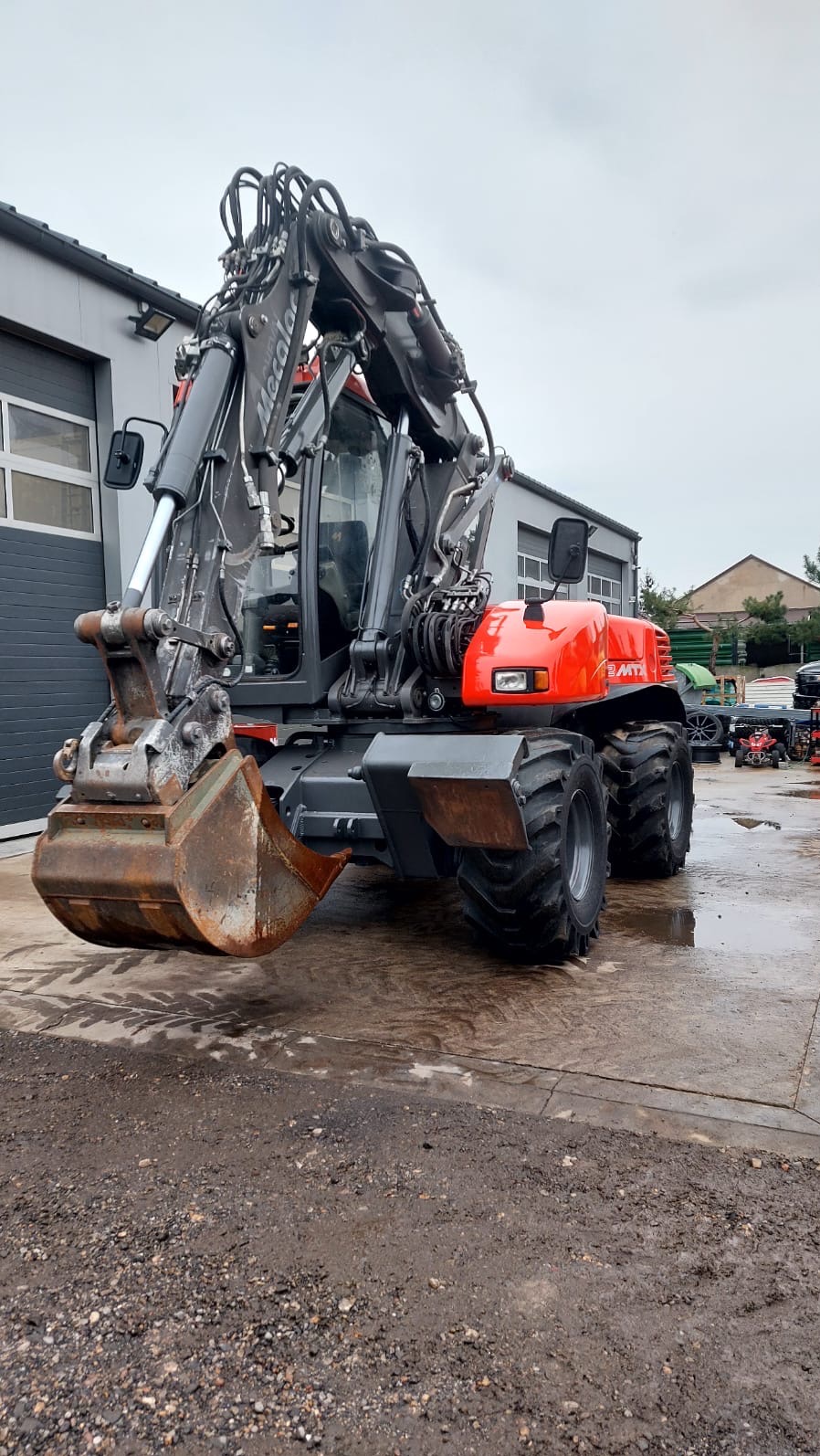 Kołowa koparka Mecalac MTX z łyżką, stojąca na mokrym placu budowy; widoczny budynek w tle.