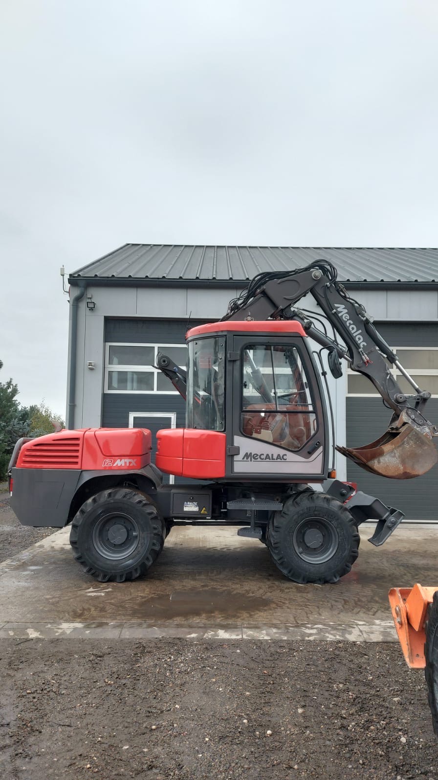Czerwono-szara koparka kołowa Mecalac 12MTX zaparkowana przed szarym budynkiem z bramą garażową, widok z boku na mokrym podłożu.