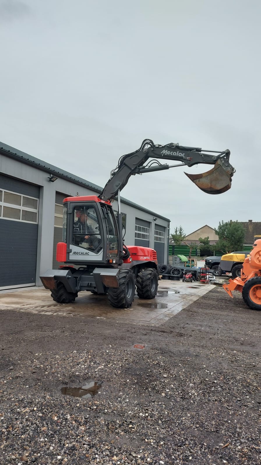 Czerwono-szara koparka kołowa Mecalac na terenie budowy, widok z przodu, operator w kabinie, lekko pochmurne niebo.