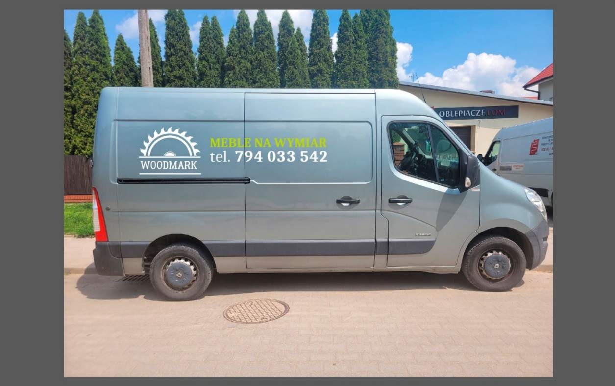 Szary samochód dostawczy z logo firmy Woodmark i napisem 'Meble na wymiar' oraz numerem telefonu, zaparkowany na ulicy w słoneczny dzień.