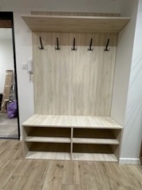 Jasna zabudowa przedpokoju z wieszakami i półkami na buty, wykonana z płyty imitującej drewno. Minimalistyczny design, widoczne elementy wykończenia wnętrza.