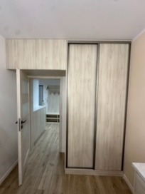 Jasna szafa wnękowa z przesuwnymi drzwiami i nadstawką, widok na wnętrze garderoby przez otwarte drzwi, drewnopodobne wykończenie, minimalistyczny design.