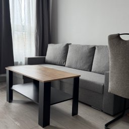 MM Cleaning Mykhailo Mykhalchuk - Jasne wnętrze pokoju dziennego: szara sofa z poduszkami, drewniany stolik kawowy z czarnymi nogami, krzesło i okno z zasłonami. Minimalistyczny, uporządkowany wystrój.