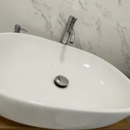 Biała, owalna umywalka nablatowa z chromowaną baterią i dozownikiem mydła na drewnianym blacie, marmurowe płytki w tle, kosz z szarej liny po prawej.