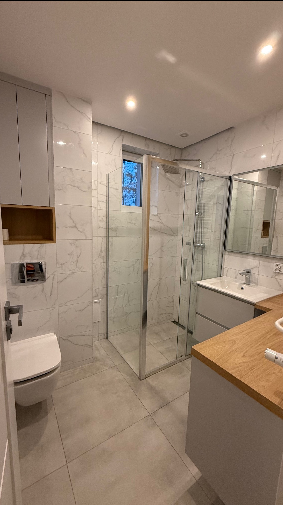 Nowoczesna łazienka z kabiną prysznicową, umywalką z drewnianym blatem i toaletą. Białe marmurowe płytki na ścianach i szare na podłodze. Widok na okno.
