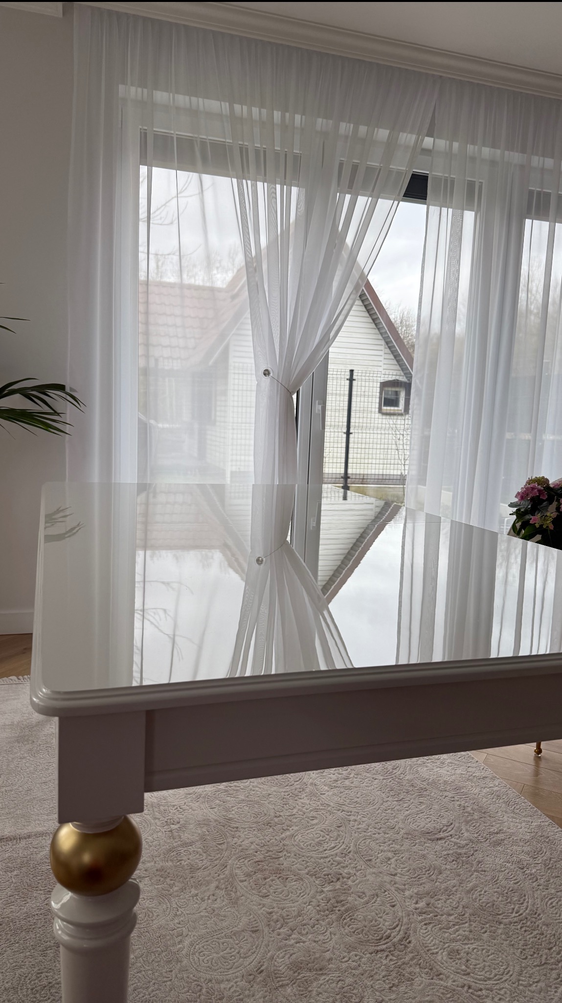 Elegancki, biały stół z połyskującym blatem i złotymi kulami u podstawy nóg, na tle okna z delikatnymi, białymi firanami i widokiem na dom.