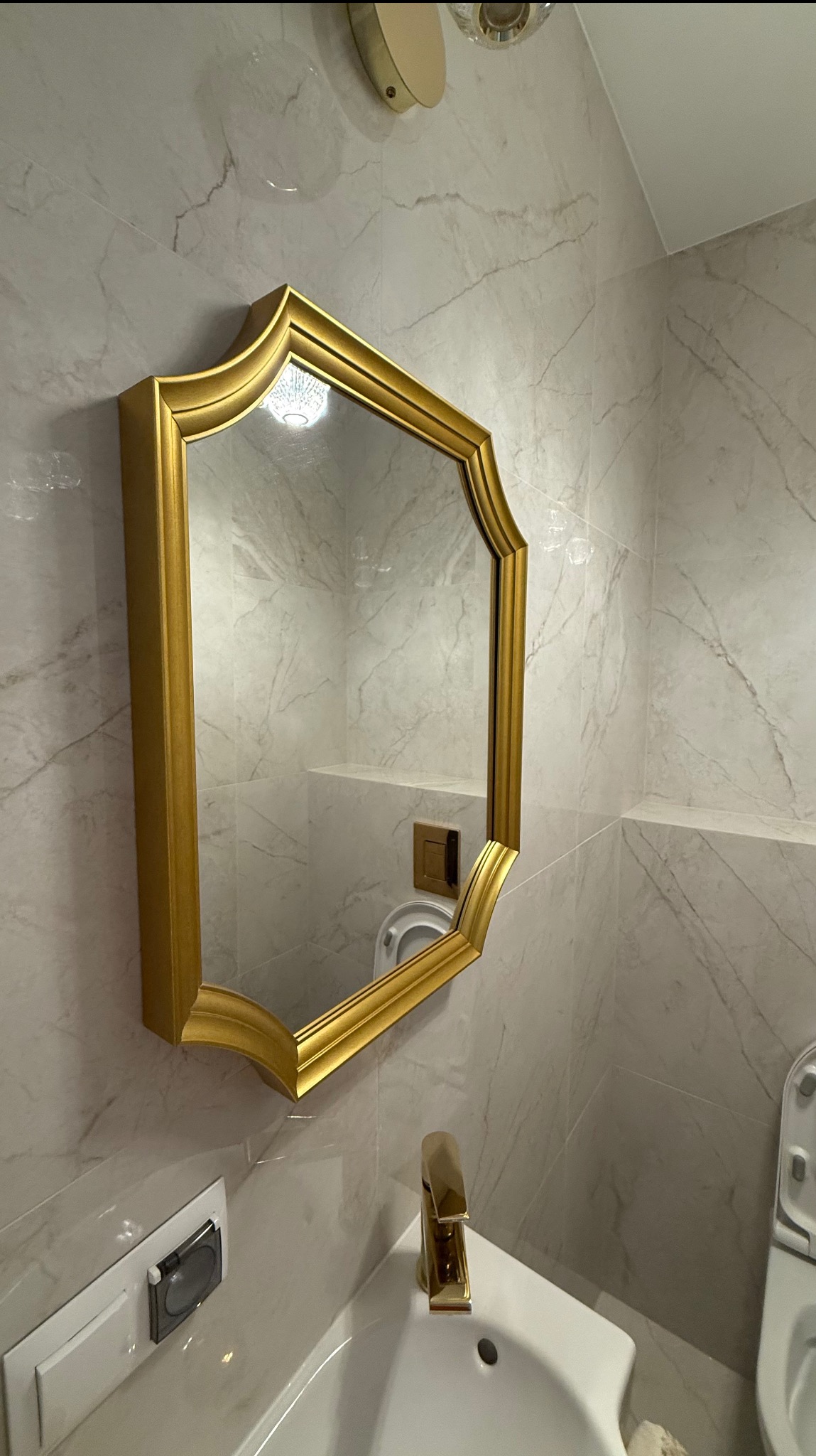 Wykończona łazienka z marmurowymi płytkami, złotą armaturą i ozdobnym lustrem. Widok na umywalkę, toaletę i fragment oświetlenia sufitowego.