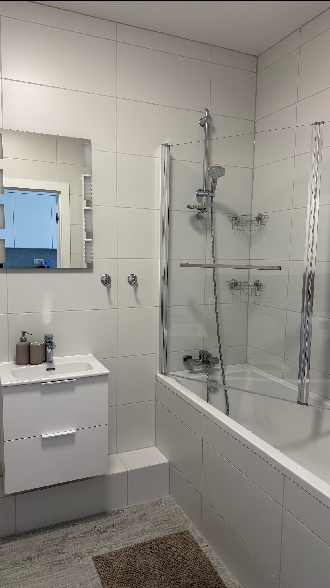 Jasna łazienka z białą armaturą, kabiną prysznicową i lustrem. Widoczne płytki na ścianach i podłodze, minimalistyczny design z akcentami w odcieniach szarości i brązu.