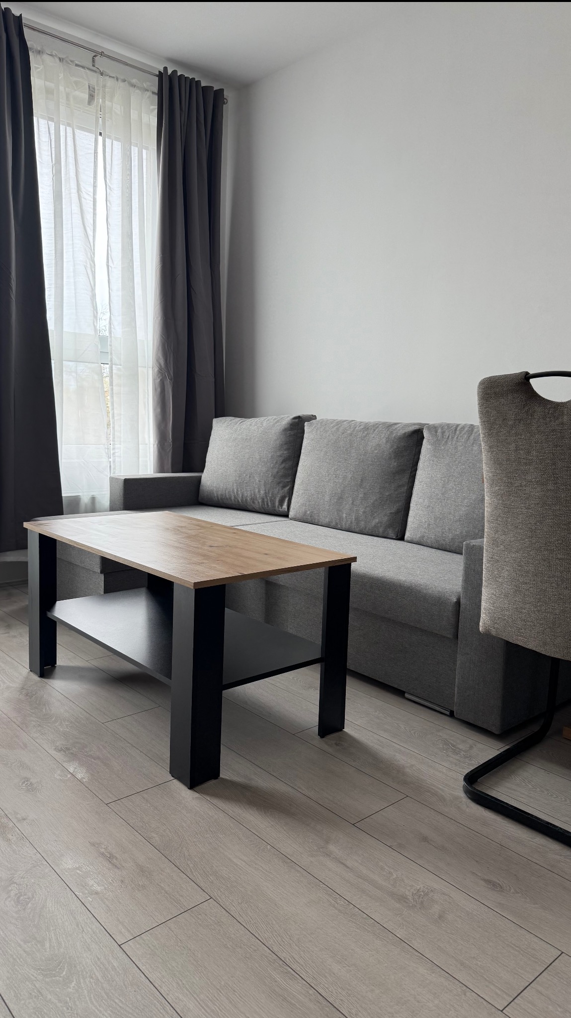 Jasne wnętrze pokoju dziennego: szara sofa z poduszkami, drewniany stolik kawowy z czarnymi nogami, krzesło i okno z zasłonami. Minimalistyczny, uporządkowany wystrój.