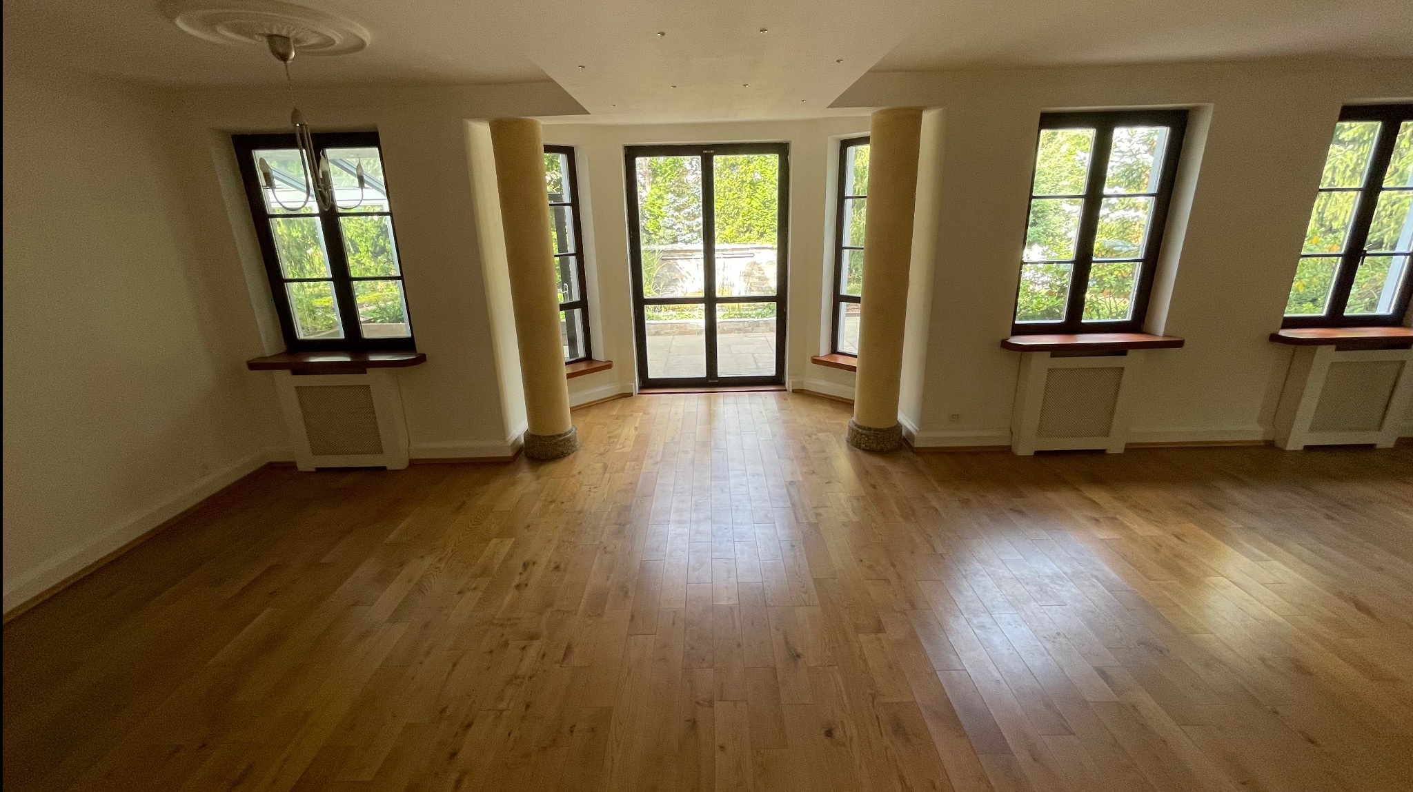 Jasne, przestronne wnętrze z parkietem, kolumnami i dużymi oknami z widokiem na zieleń. Okna z ciemnymi ramami i drewnianymi parapetami. Wnętrze po sprzątaniu.