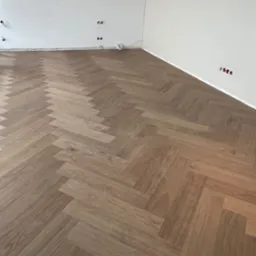 Jasnobrązowa podłoga ułożona w jodełkę francuską w nowym pomieszczeniu z białymi ścianami i widocznymi gniazdkami elektrycznymi bez osprzętu.