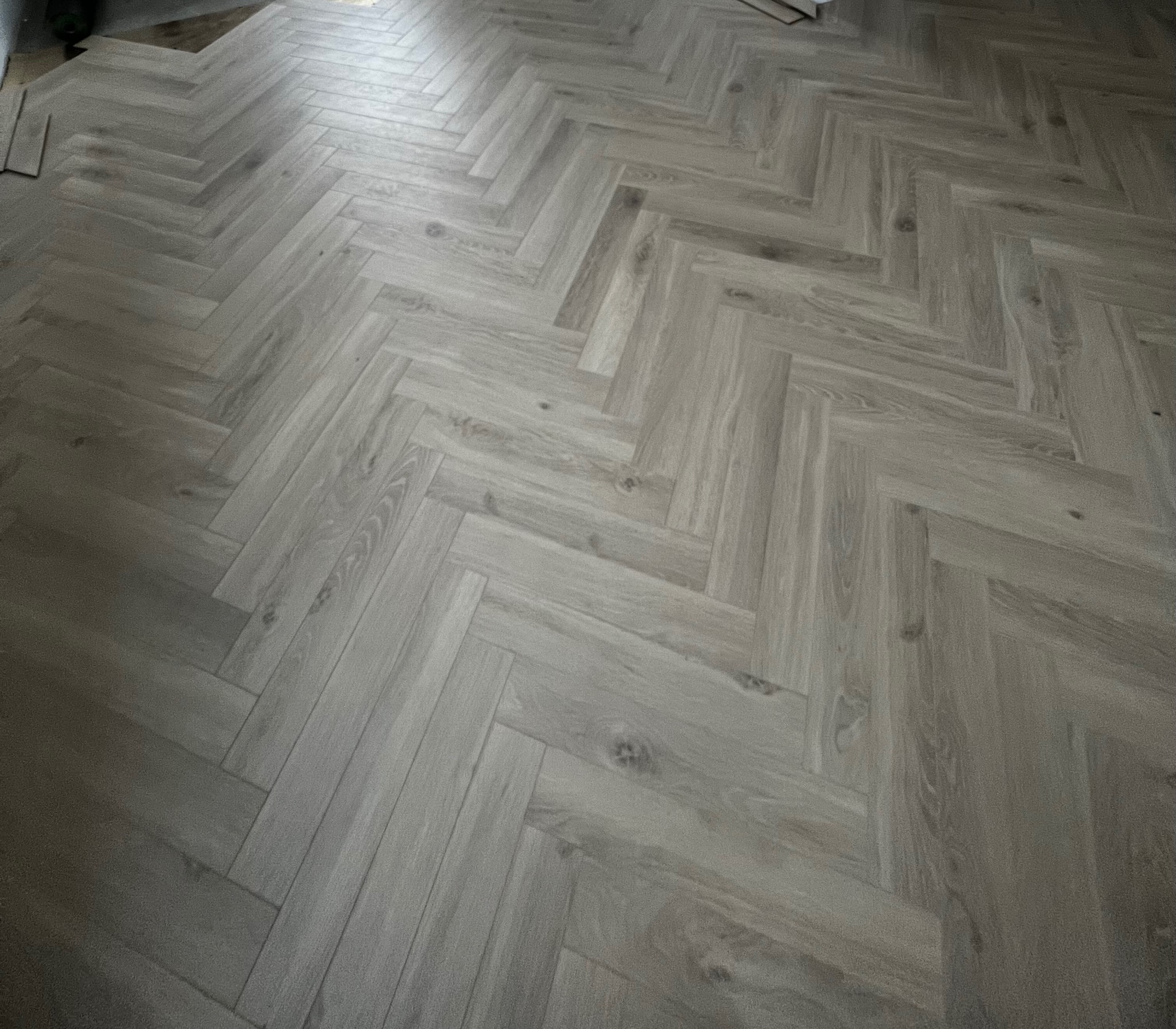 Ułożona pod kątem jodełki podłoga z paneli laminowanych w odcieniach szarości, z widocznymi detalami słojów drewna i fragmentami niezakończonej instalacji w rogu.