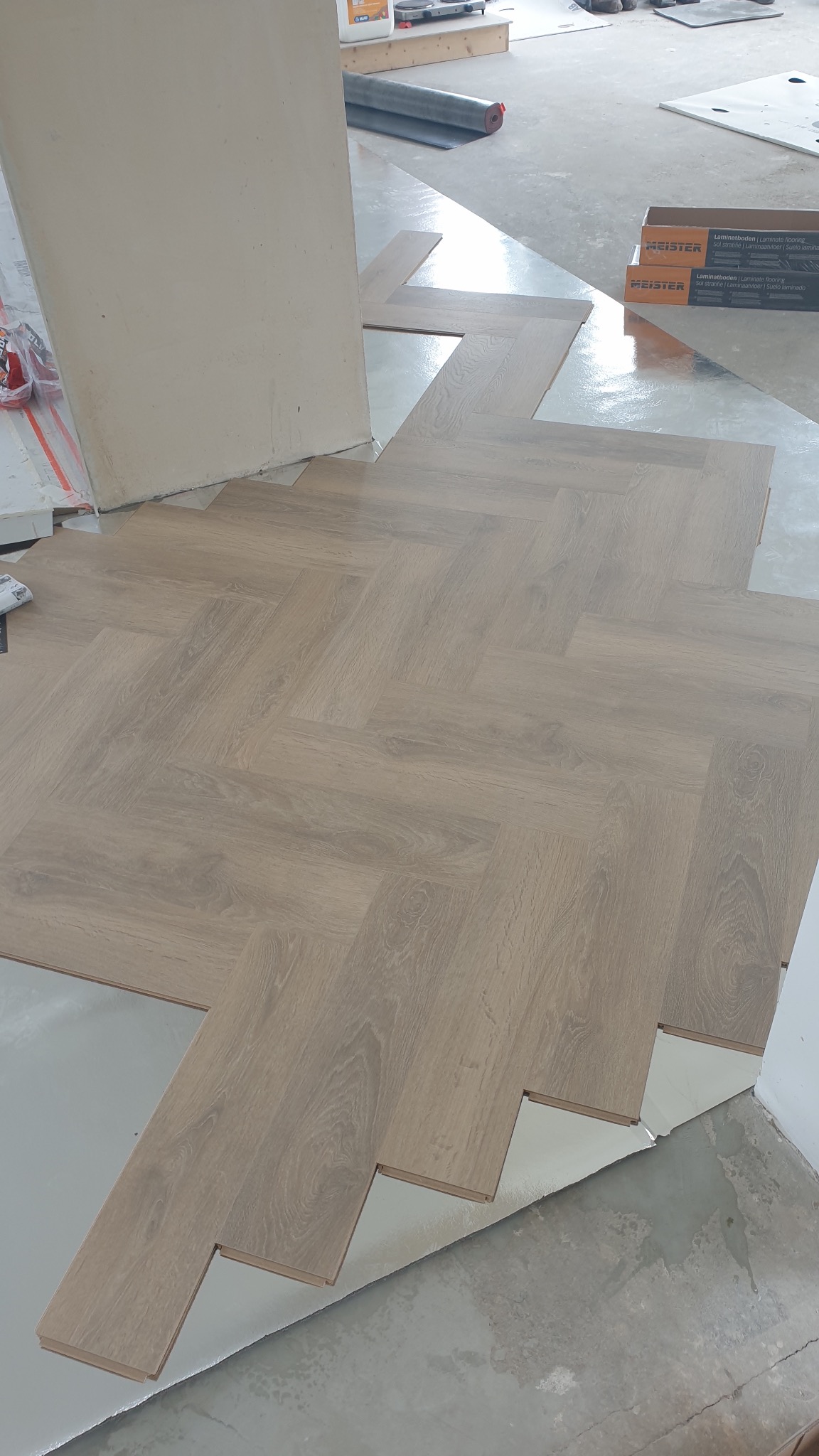 Fragment podłogi z ułożonym wzorem jodełki z paneli laminowanych, widoczne pudełka po panelach Meister, podkład pod panele i fragment surowej ściany.