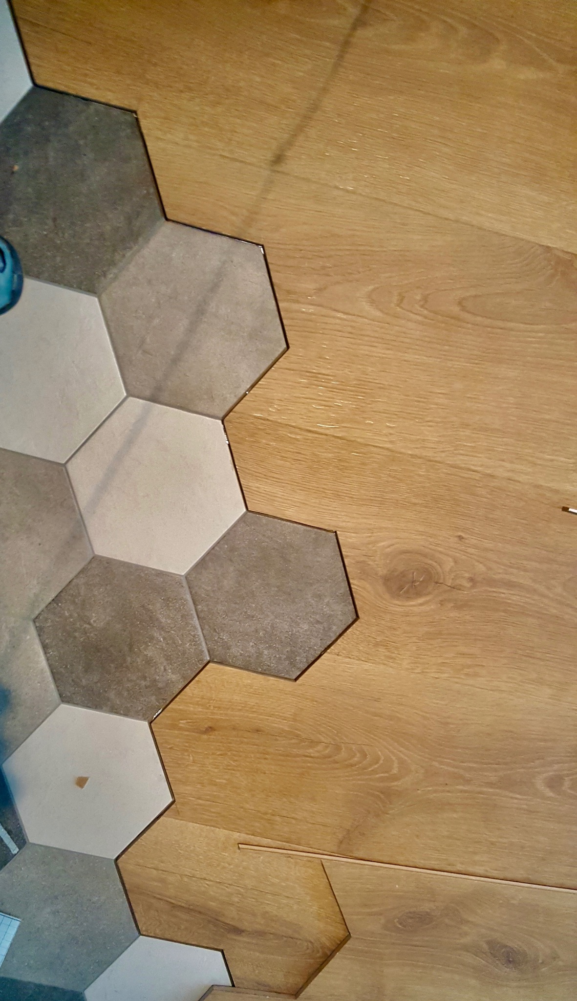 Fragment podłogi z połączeniem heksagonalnych płytek w odcieniach szarości i beżu oraz paneli imitujących drewno, widoczne drobne elementy montażowe.