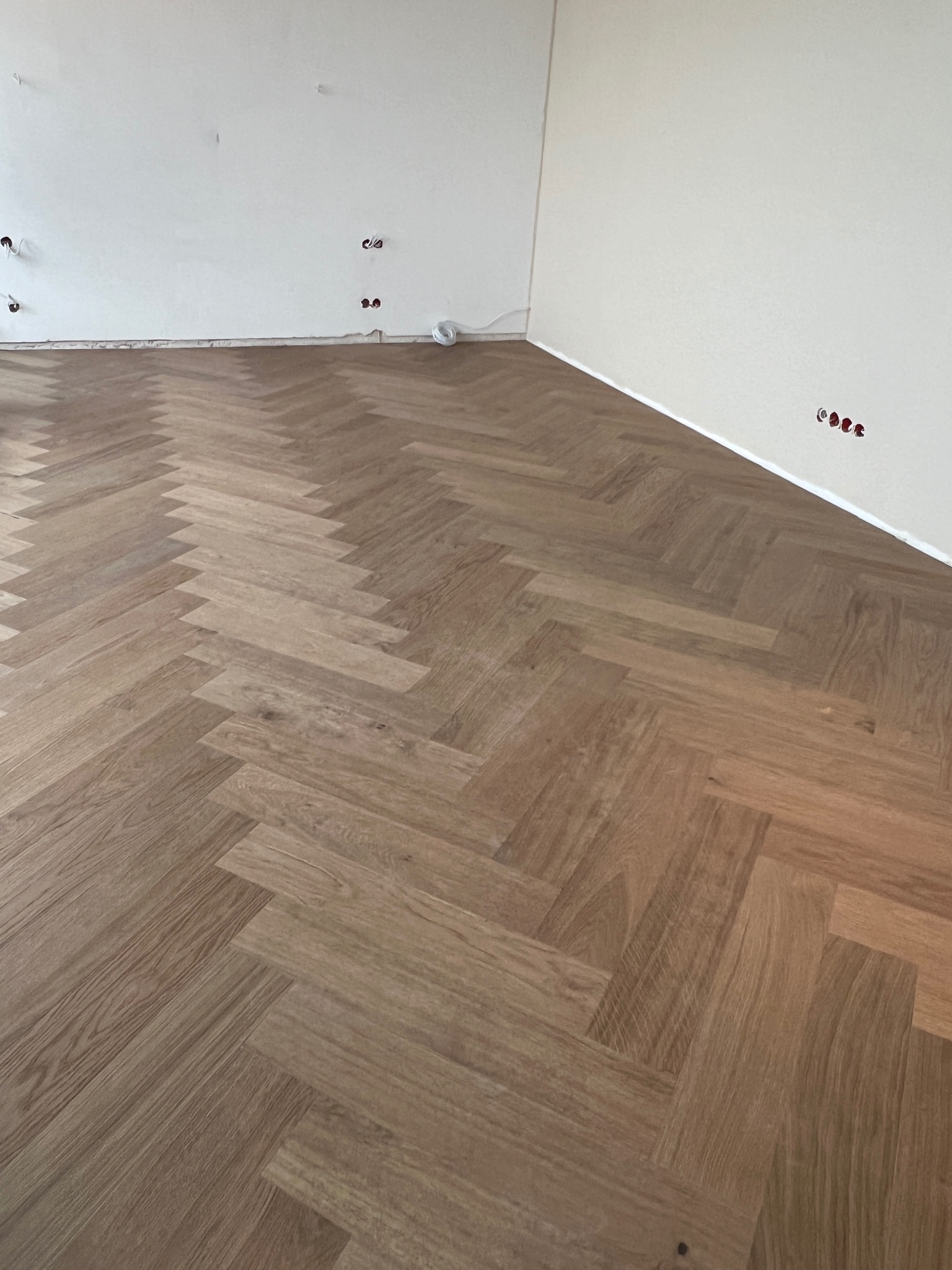 Jasnobrązowa podłoga ułożona w jodełkę francuską w nowym pomieszczeniu z białymi ścianami i widocznymi gniazdkami elektrycznymi bez osprzętu.