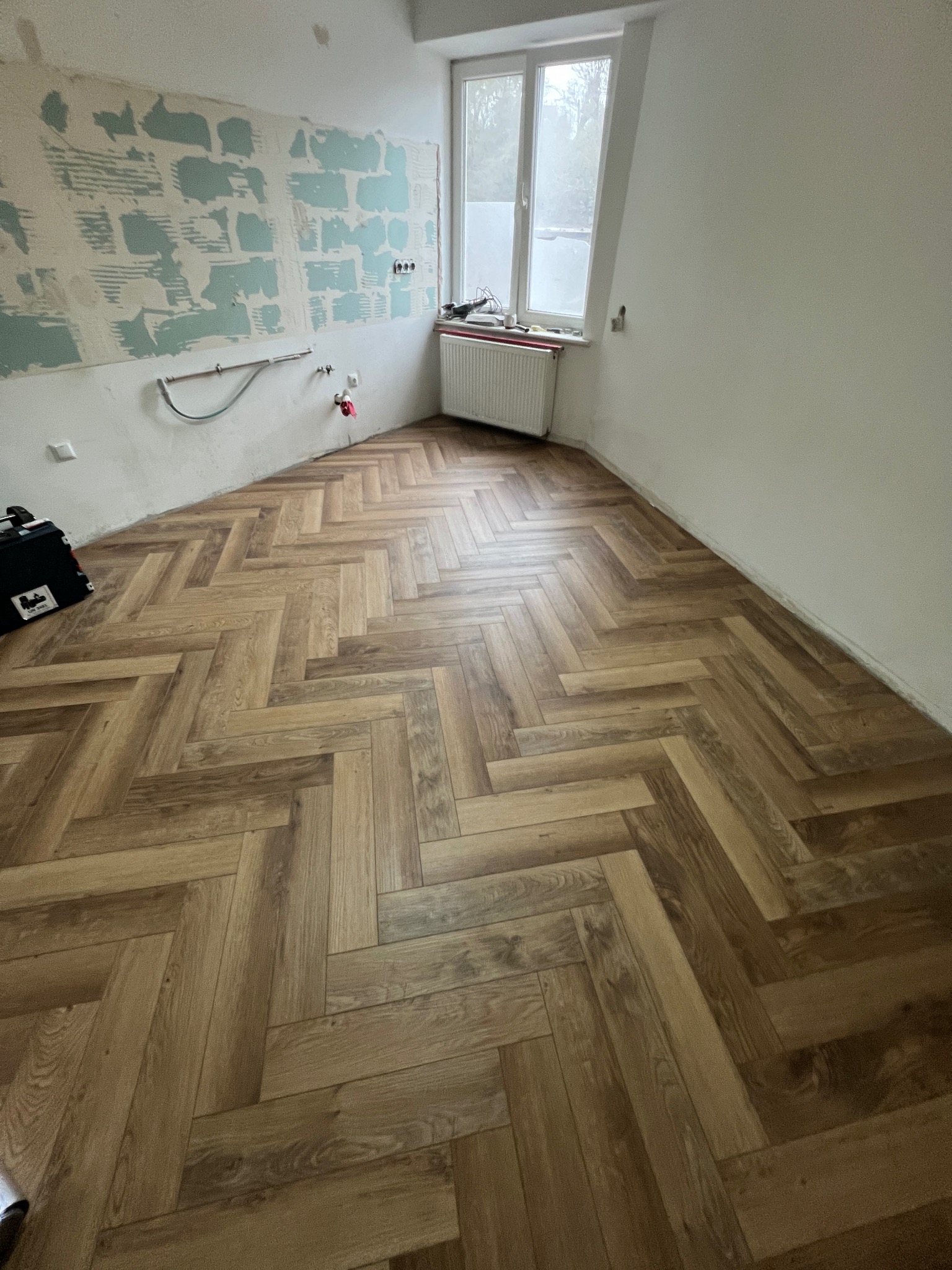 Ukończone układanie drewnianych paneli podłogowych w jodełkę w pokoju z przygotowaną ścianą do malowania i oknem.