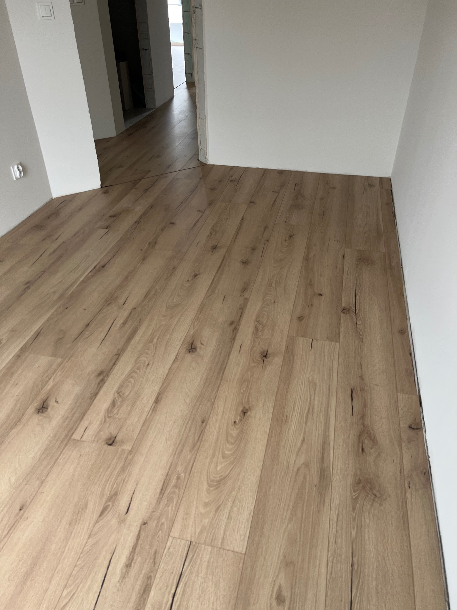 Ułożone panele podłogowe o jasnym odcieniu drewna, widoczne przejście do kolejnego pomieszczenia z identyczną podłogą, białe ściany i listwy przypodłogowe.