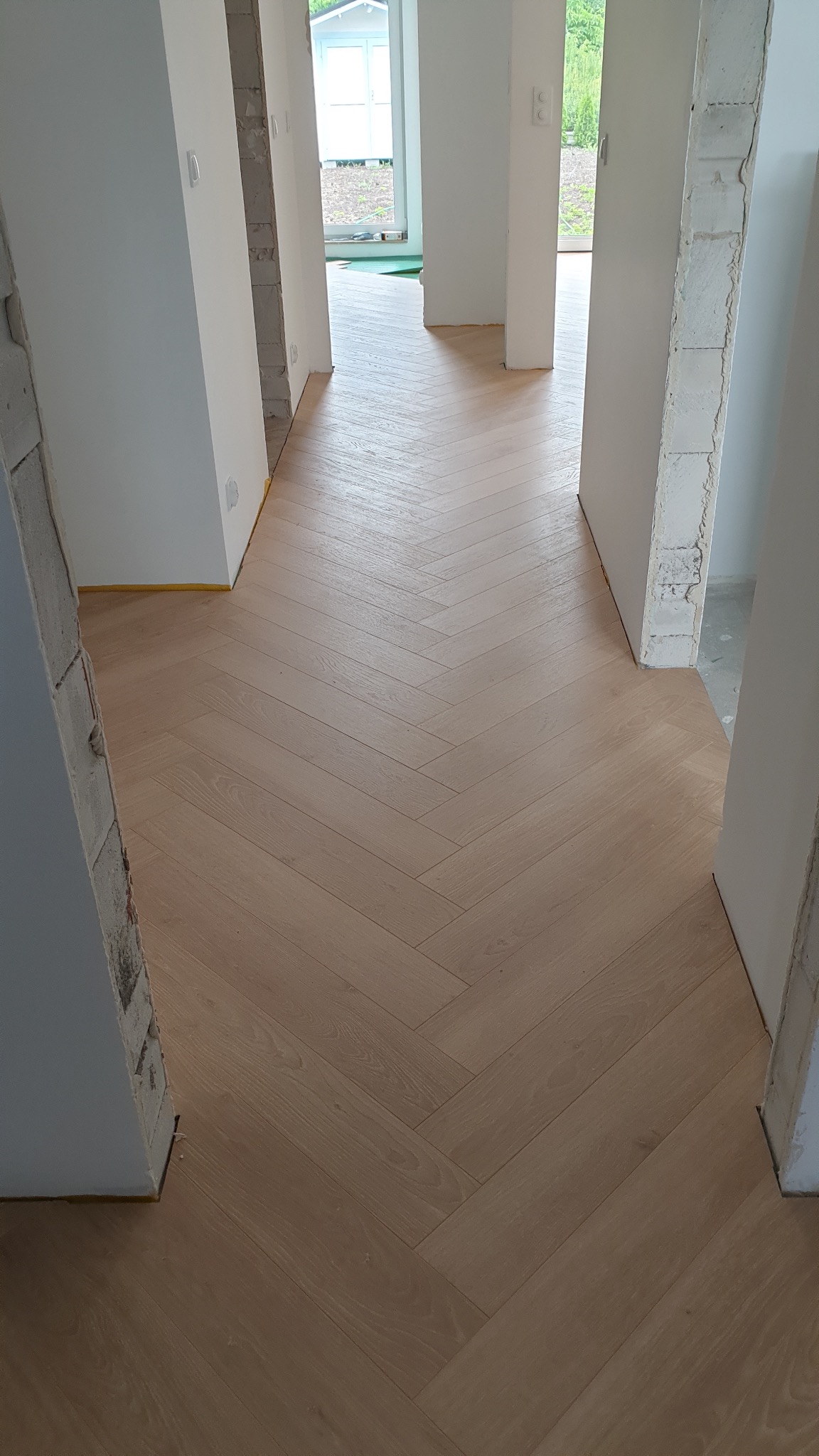 Jasne panele ułożone we wzór jodełki w korytarzu z widocznymi surowymi fragmentami ścian i żółtą taśmą zabezpieczającą.