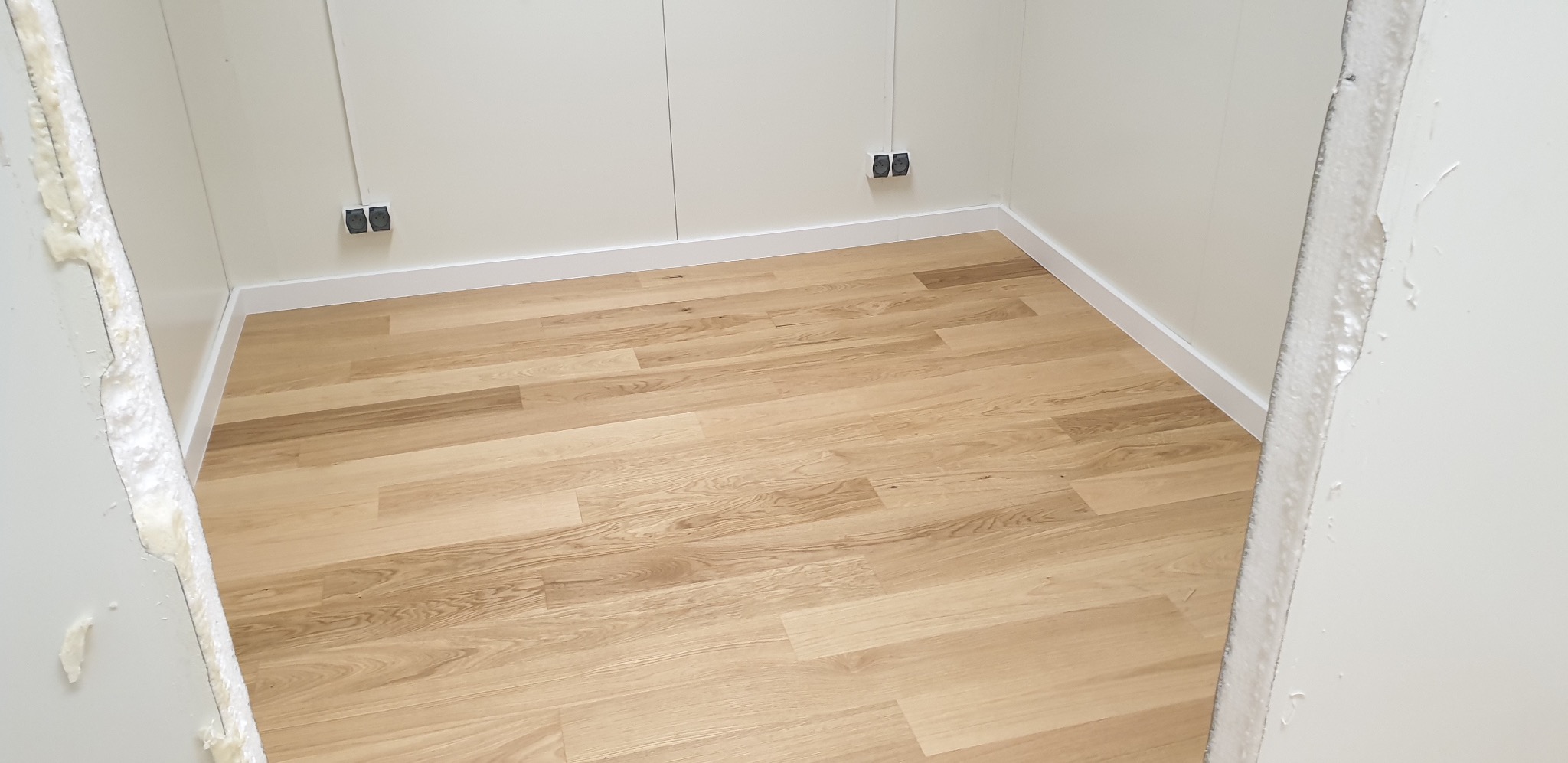 Jasne panele podłogowe ułożone w rogu pomieszczenia z białymi ścianami i listwami przypodłogowymi, widoczne gniazdka elektryczne i surowa krawędź ściany z pianką montażową.