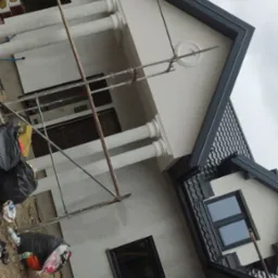 Niedokończona elewacja domu z widocznymi rusztowaniami, workami na śmieci i żółtym wężem wentylacyjnym, w tle pochmurne niebo.